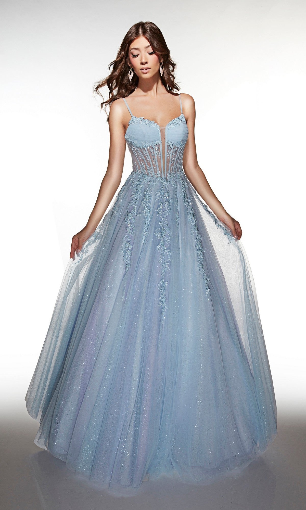 A woman stands against a white background wearing the Alyce 62061 long prom dress in light blue/lilac, featuring a floor-length skirt, lace corset bodice, delicate spaghetti straps, and floral embellishments.